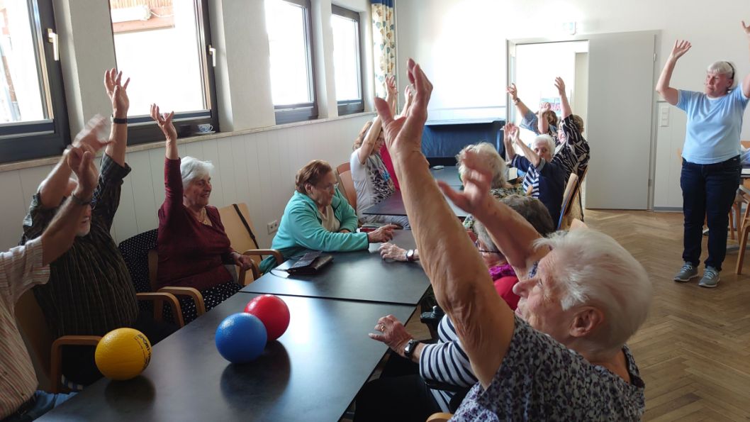 Das Bild zeigt einen Teil der Bewegungsübungen mit Schulterkreisen von vorn nach hinten und von hinten nach vorn; Kopf im Wechsel nach links und rechts drehen; Arme angewinkelt vor dem Körper halten und öffnen, schließen; mit der rechten Hand ans linke Knie fassen, dann mit der linken Hand ans rechte Knie… Bild: Sportkreis GG