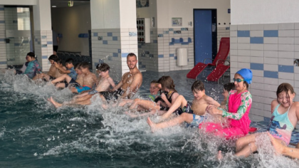Bewegungskoordinator Marius Hamel zeigt den Kindern eine Übung zur Wassergewöhnung: Mit den Füßen ins Becken strampeln, Spritzwasser erleben und spielerisch Wasserwiderstand, Strömungen und das Element Wasser kennenlerne.                Bild: Sportkreis Waldeck-Frankenberg